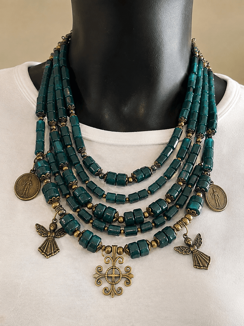 Emerald Benediction necklace worn at the neck, with layered teal beads, brass spacers, angel charms, and a cross pendant